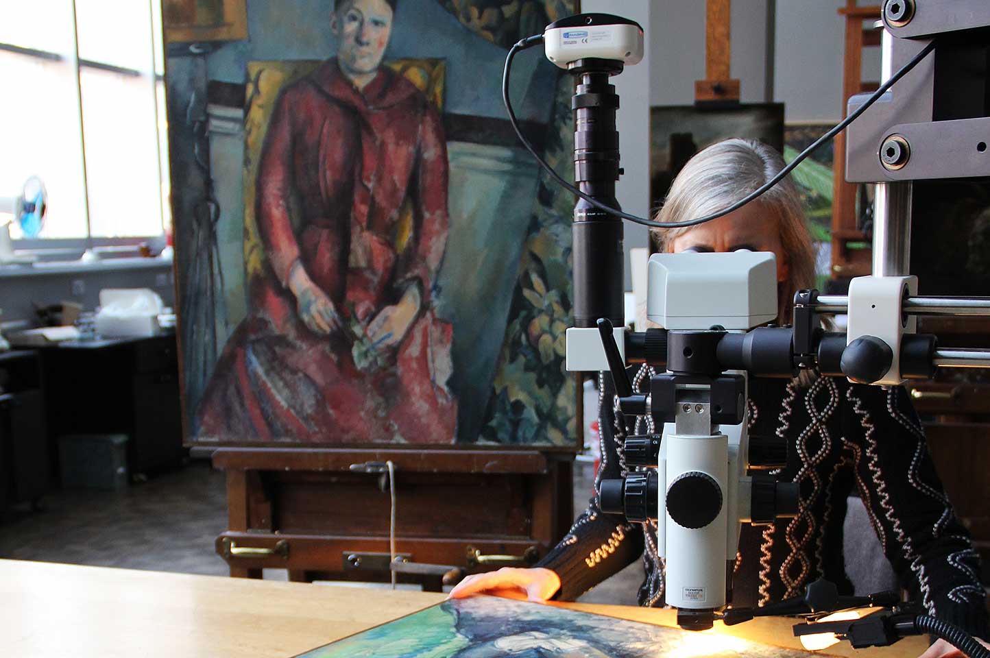 Conservator looking through a microscope at a Cezanne painting on a table with another Cezanne painting displayed on an easel in the background